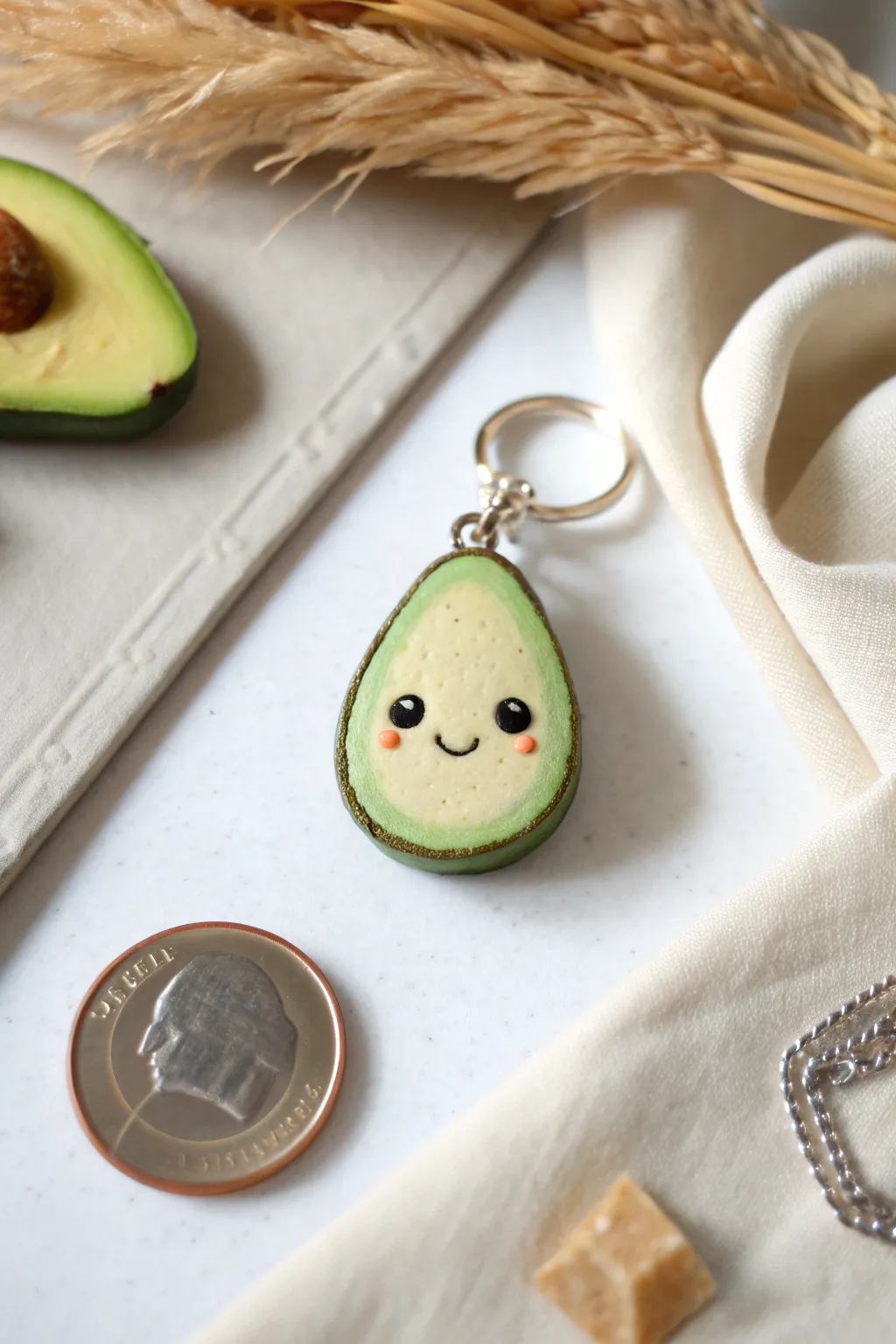 Fingernail-size smiling avocado clay charm with a tiny loop, styled minimally with a coin for scale