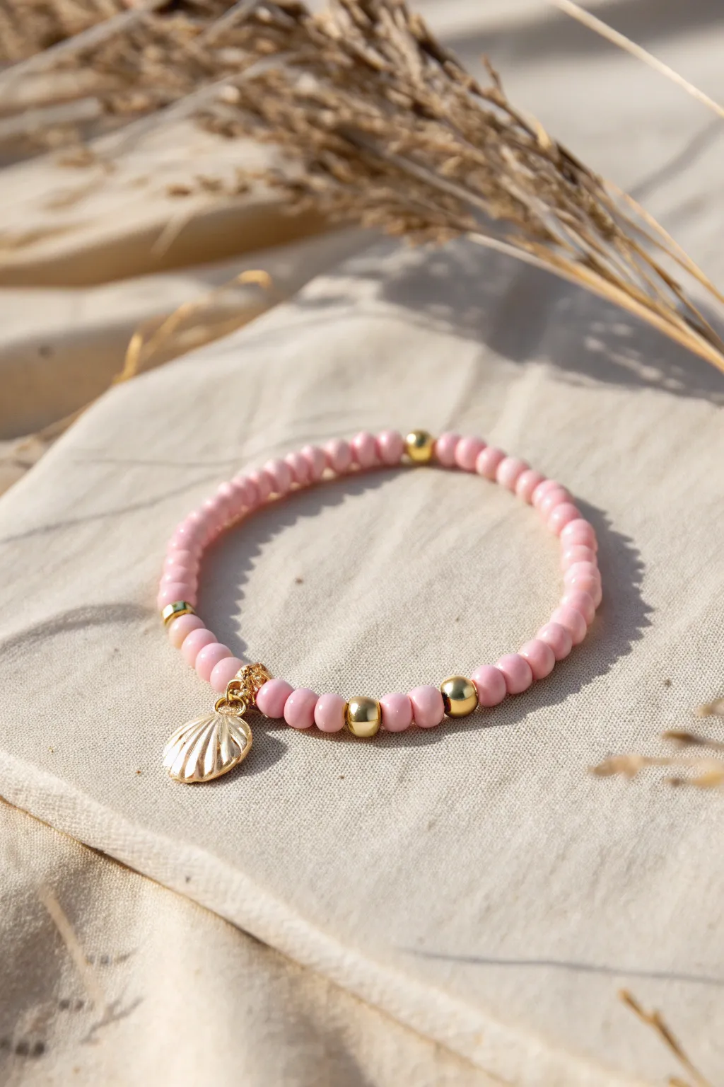 Pink beach-prep heishi bracelet with shell accent and gold spacers on sandy linen.