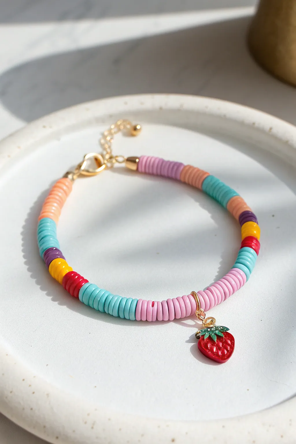 Tiny fruit charm pop on a colorful heishi clay bead bracelet, clean minimalist flat lay.