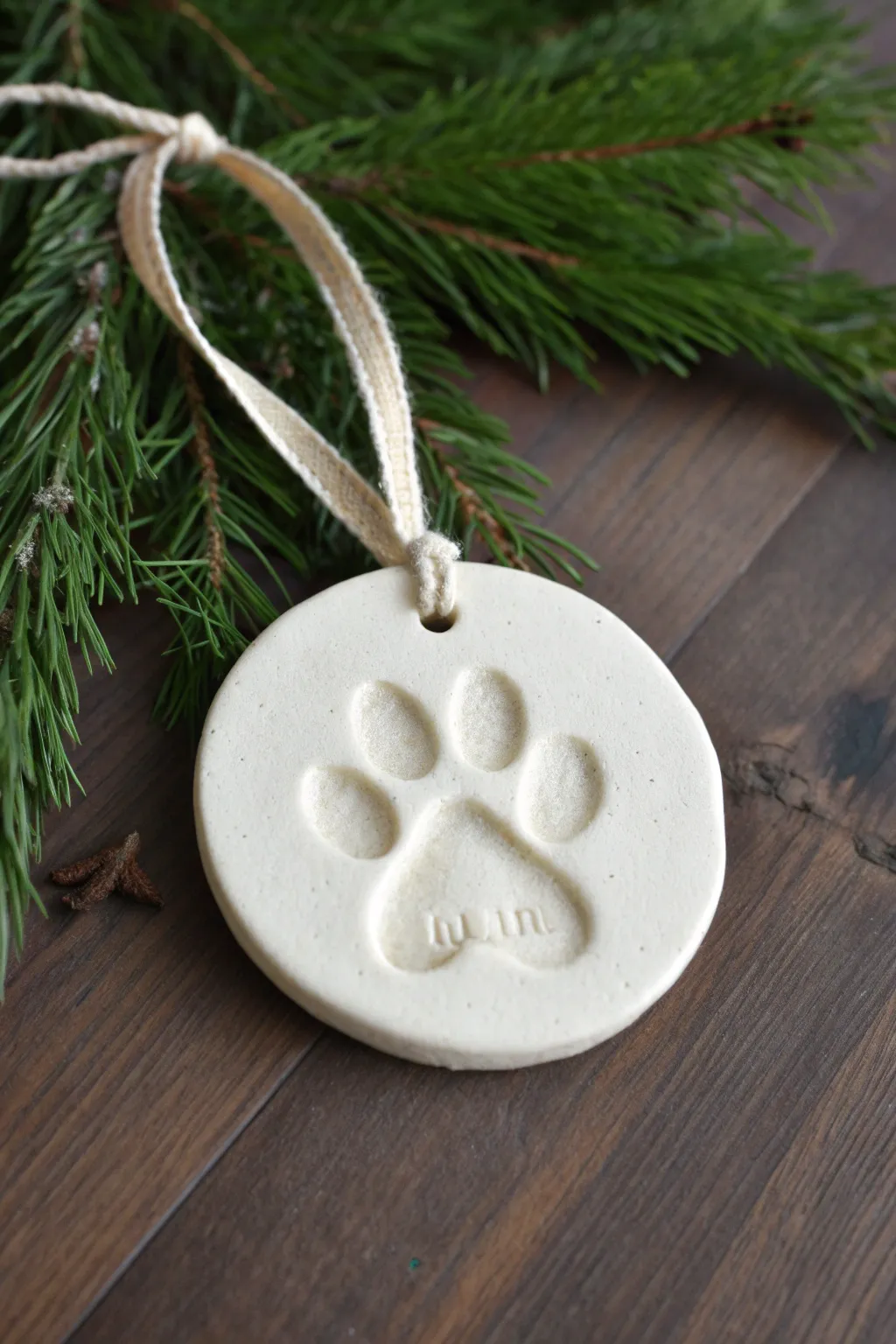 Sweet paw print holiday keepsake ornament in clay, minimalist style with linen ribbon and greenery.