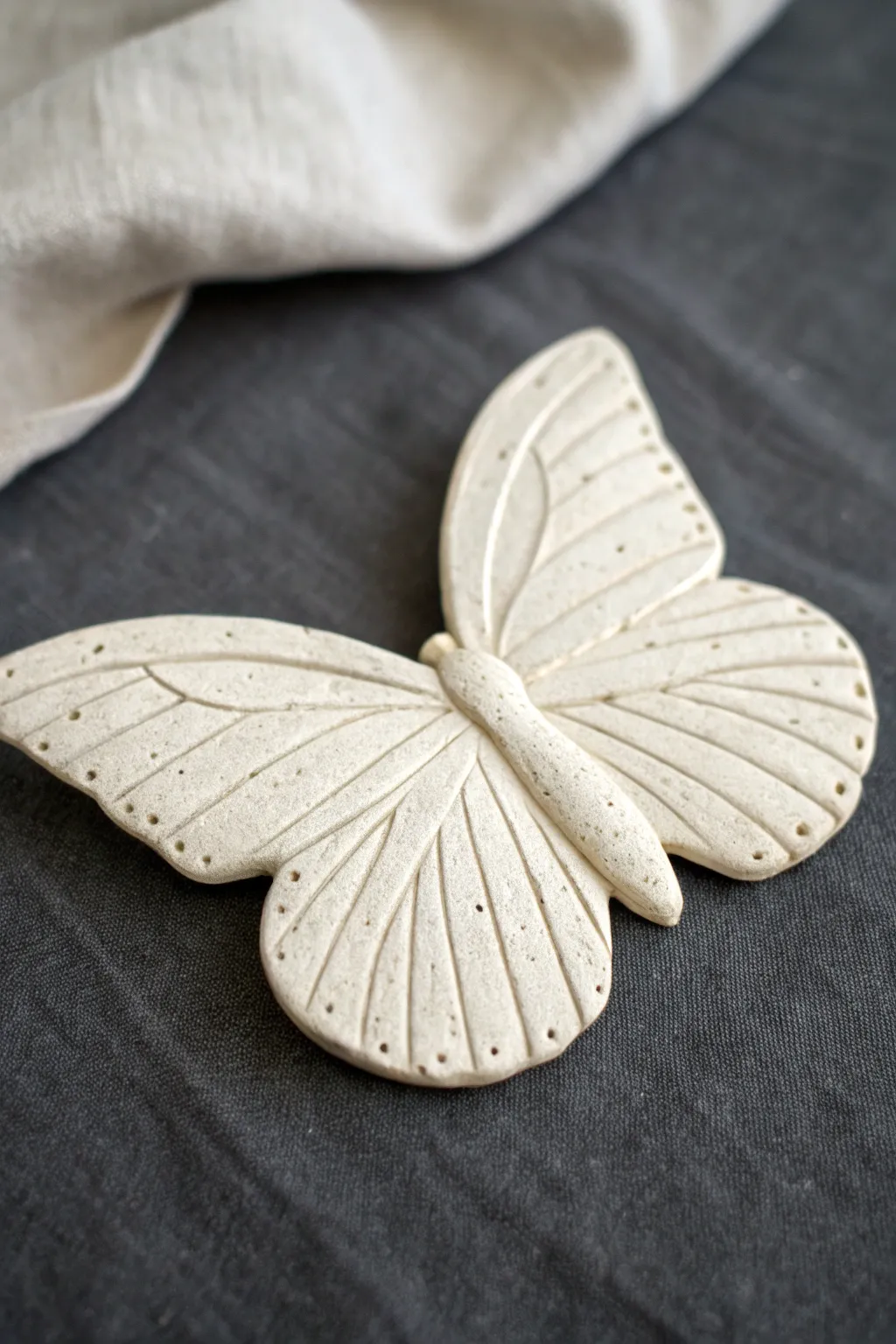 Matte clay butterfly with carved wing veins, softly lit on a dark linen background.