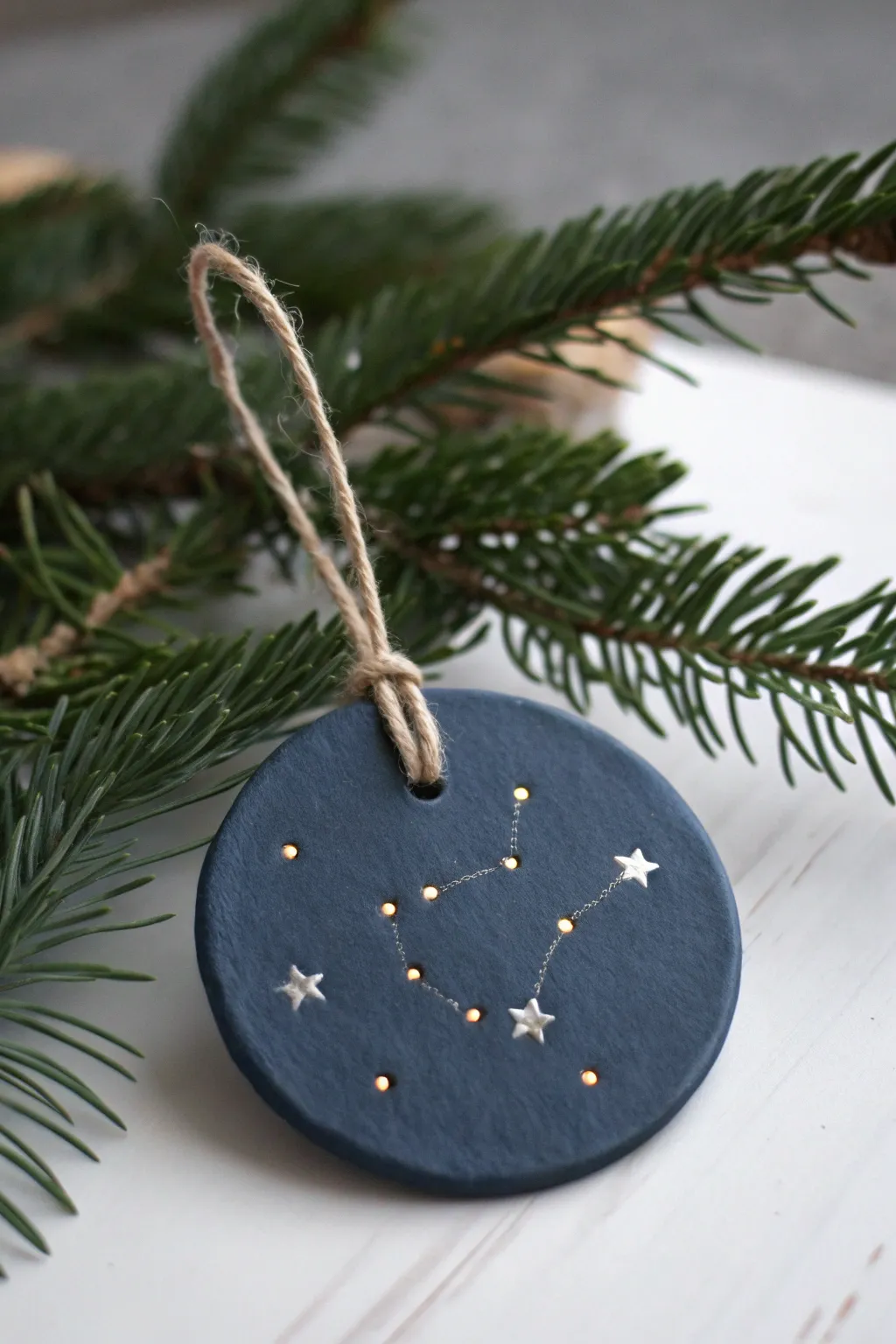 Midnight clay ornament with metallic constellation dots, softly glowing on evergreen needles.