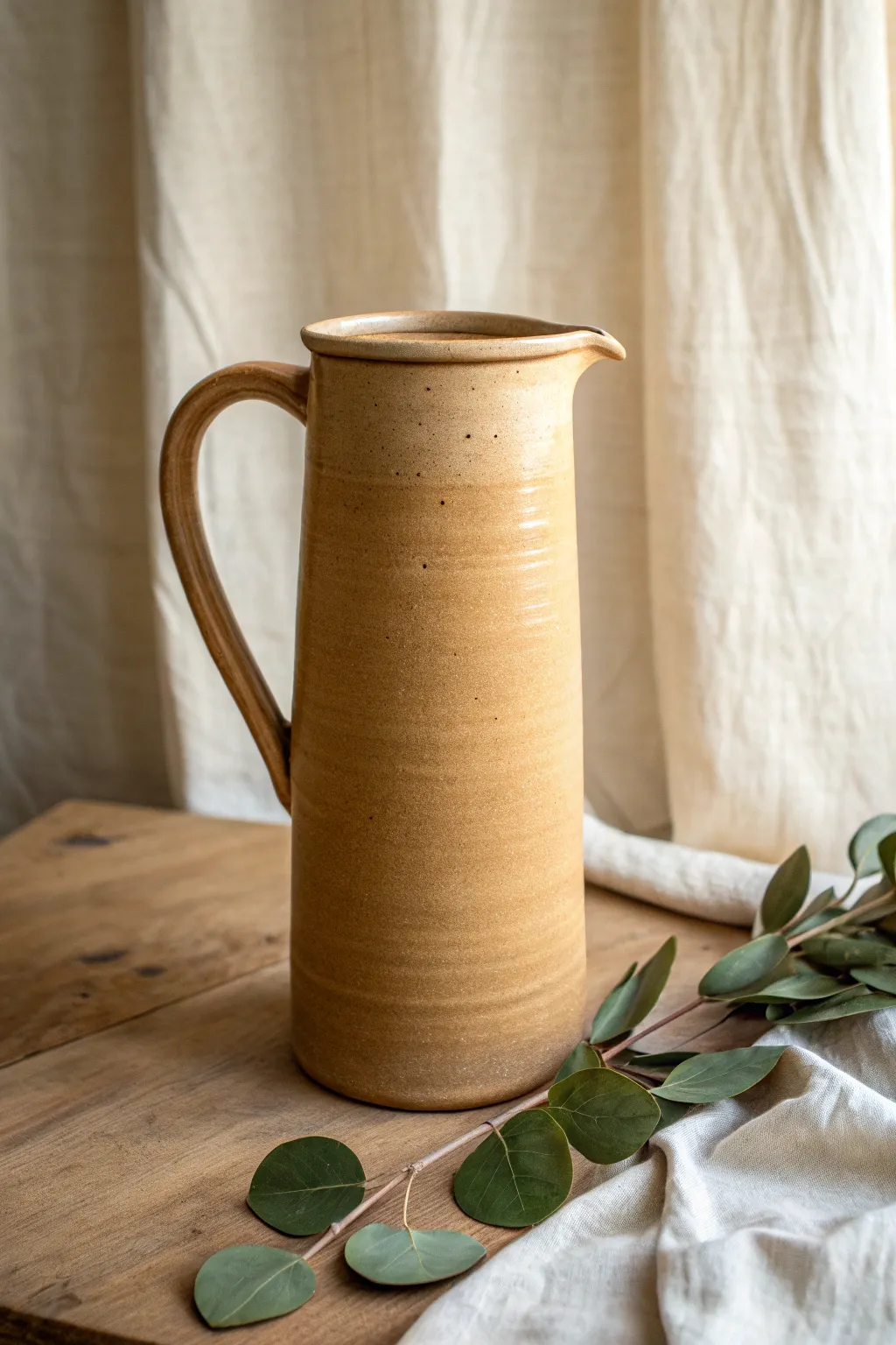 Tall minimalist clay cylinder pitcher with a narrow pour, styled with a single leafy stem.
