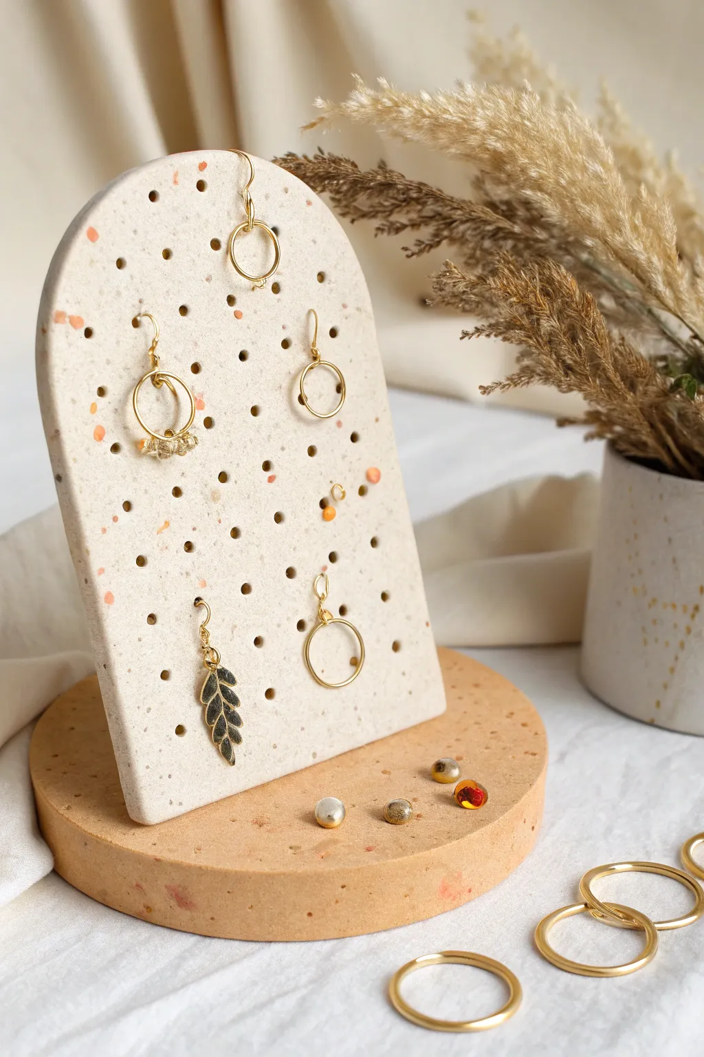 Round terrazzo clay earring board with neat holes, holding simple studs and hoops in warm minimal style