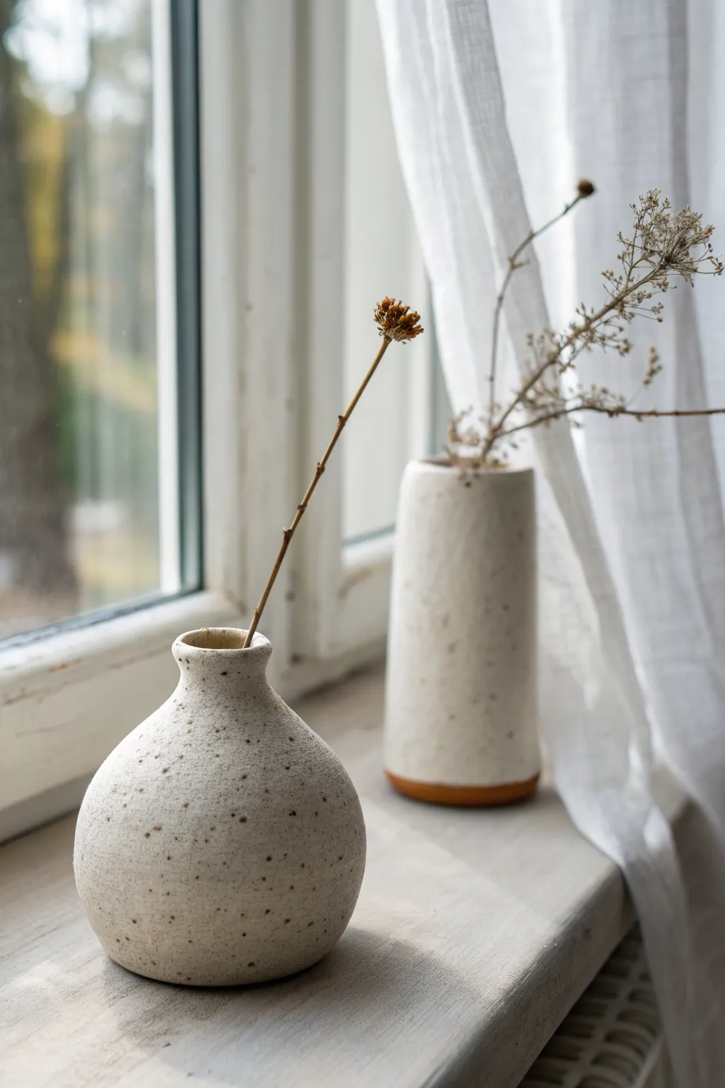 Two mini clay bud vases in soft window light, minimalist shapes with a single dried stem.