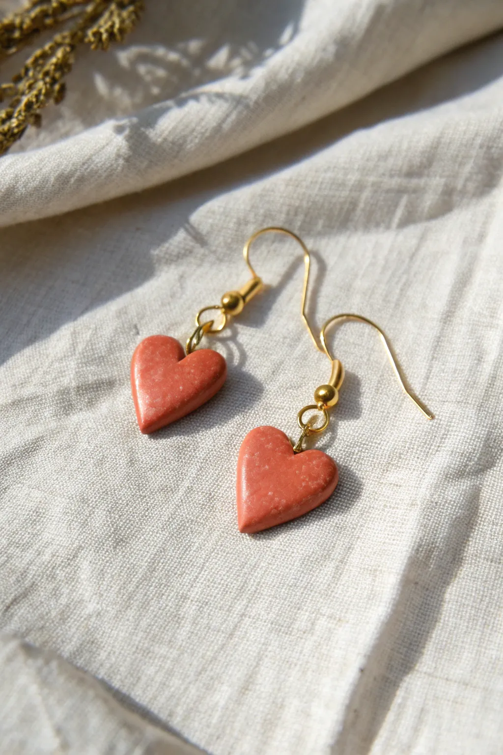 Tiny heart drop earrings in bold terracotta red, softly sealed and styled on neutral linen.