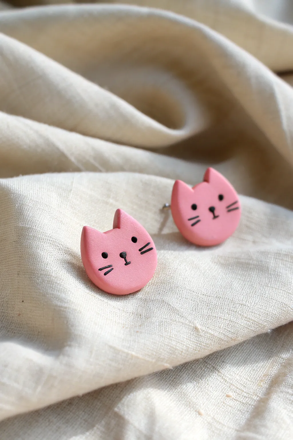Pink cat face stud earrings with rosy cheeks, neatly styled on linen for a clean boho look.