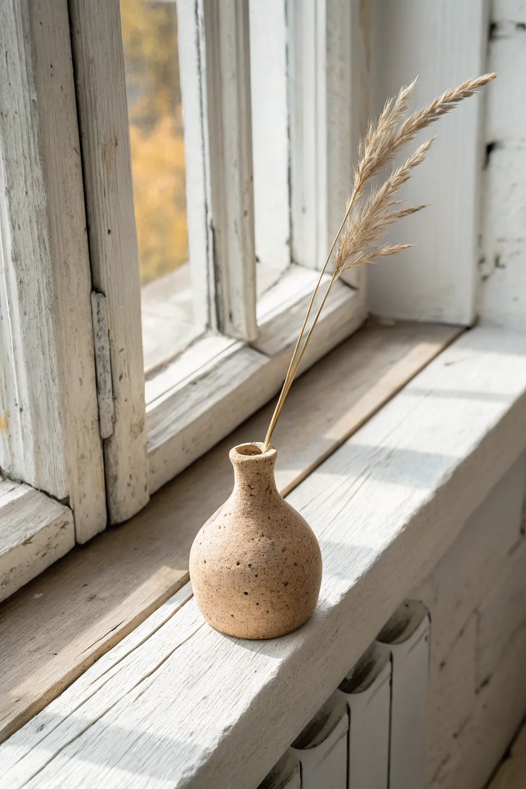 Small handmade bud vase in warm clay, styled with a single dried stem in soft window light.