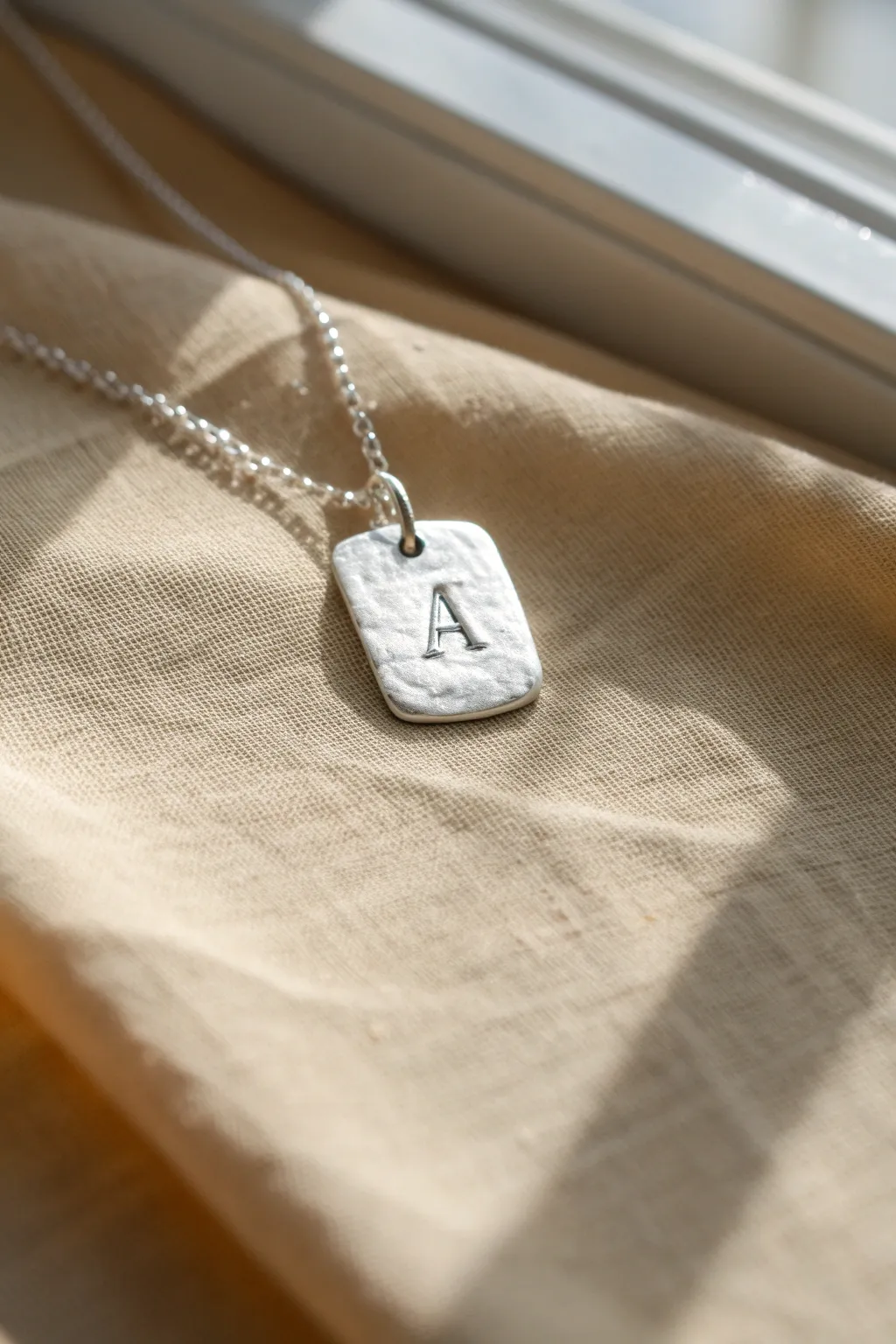 Minimal silver clay initial tag charm with a soft hammered texture, styled in calm Nordic tones.