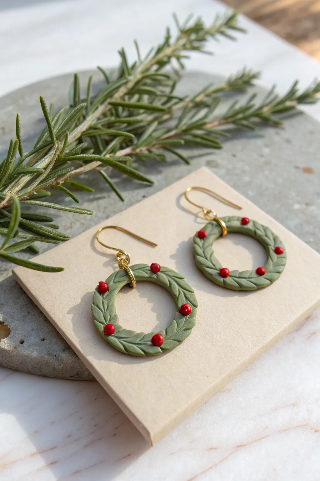 Chunky green clay wreath earrings with tiny red berries, styled with rosemary on a neutral card.