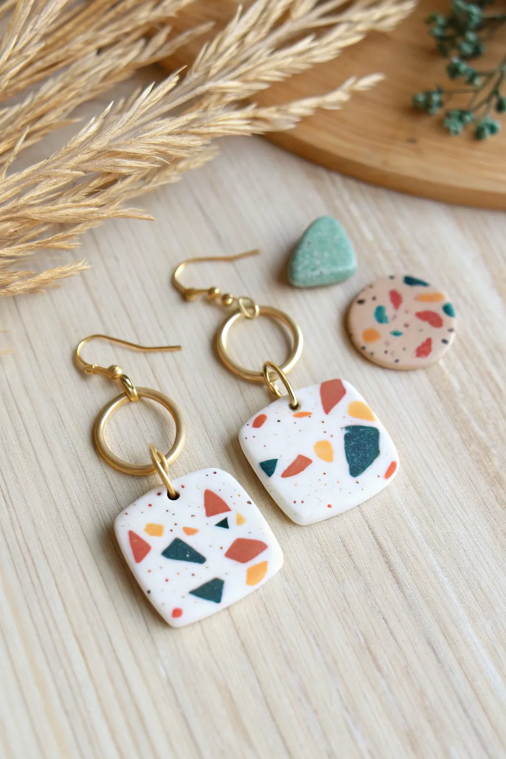 Terrazzo-speckled air dry clay tile earrings with simple gold hoops on pale wood.