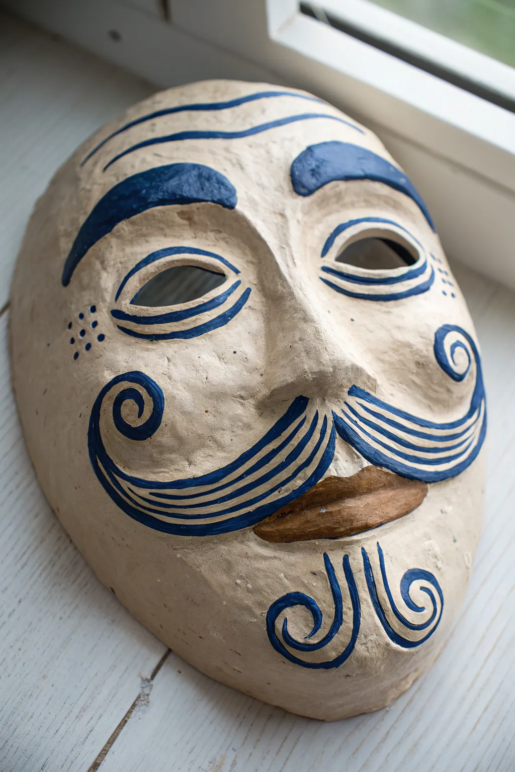Playful coil-brow and mustache clay mask in earthy terracotta with bold indigo accents.