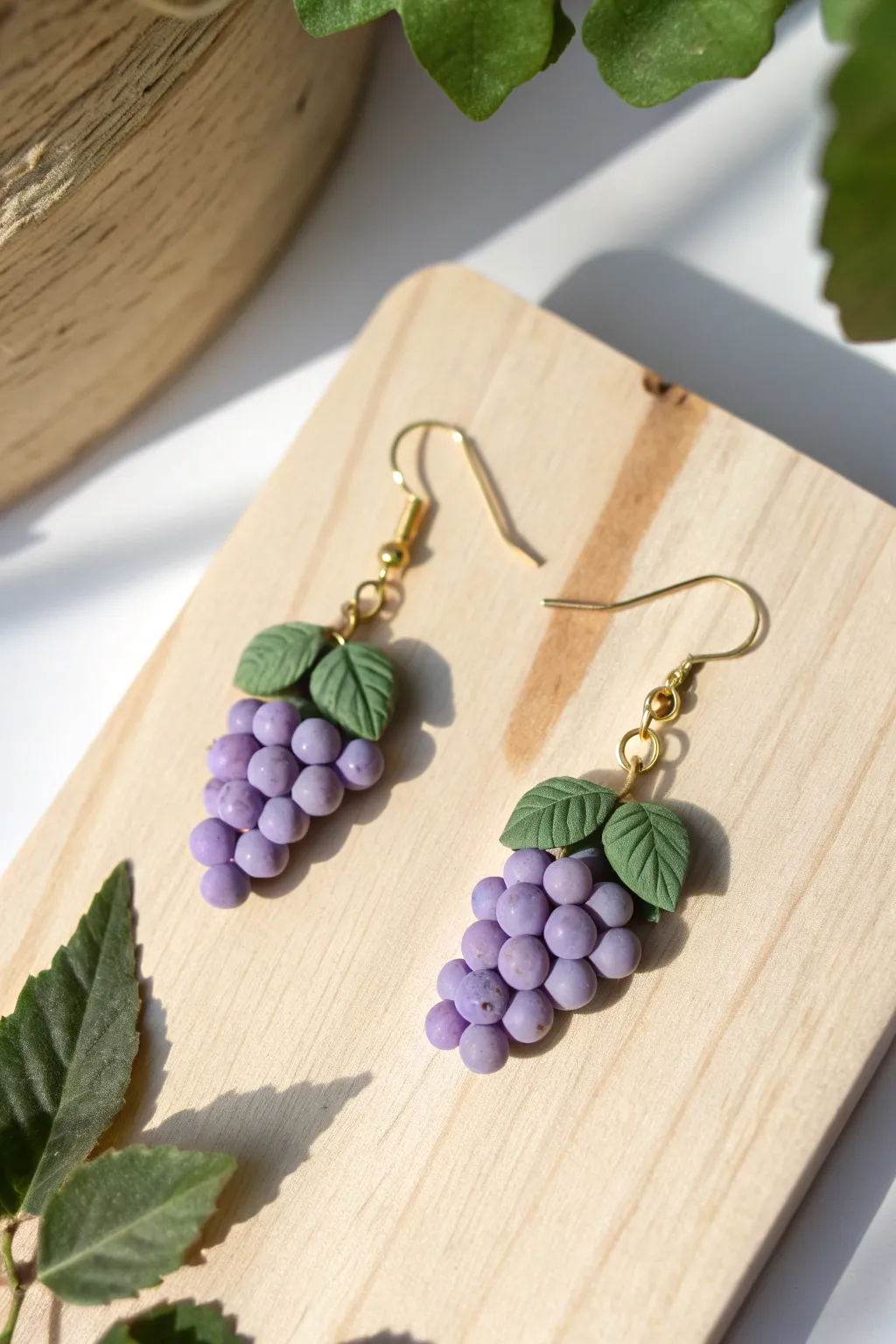 Grape cluster clay earrings in deep purple with a tiny green leaf, hanging cleanly on light wood.